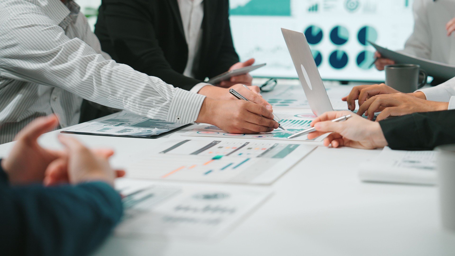 Group of business professionals collaborating on financial reports and charts during a meeting in a modern office setting with laptops and documents. SACTR